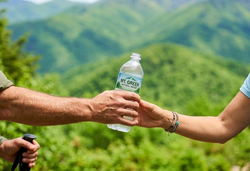 登山の途中で、男性が女性にペットボトルの水を優しく手渡している手元の写真。背景には美しい新緑の山々が広がっている健康的なイメージ。